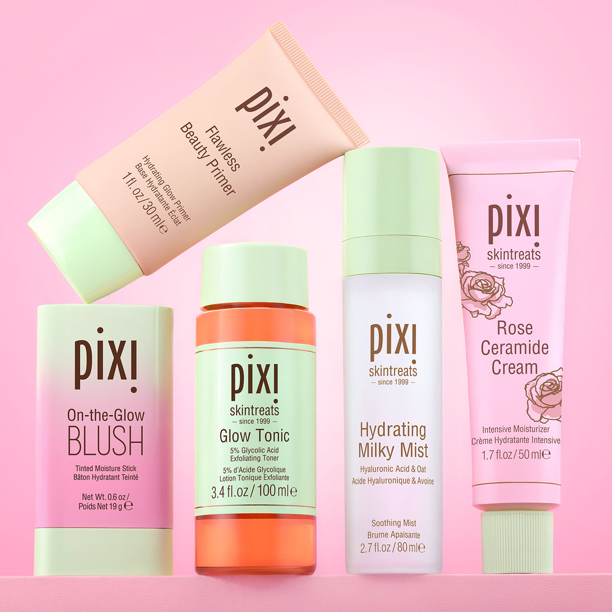 Six Pixi skincare and cosmetic products arranged together on a pink background. The products include Pixi Flawless Beauty Primer, Pixi On-the-Glow Blush, Pixi Glow Tonic, Pixi Hydrating Milky Mist, Pixi Rose Ceramide Cream. On a pink background.