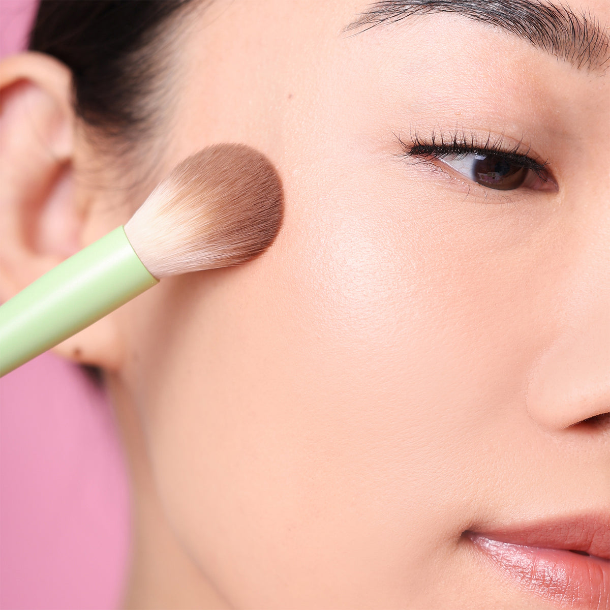 Close-up of a person applying makeup with Pixi Glow Brush on a blurred pink background