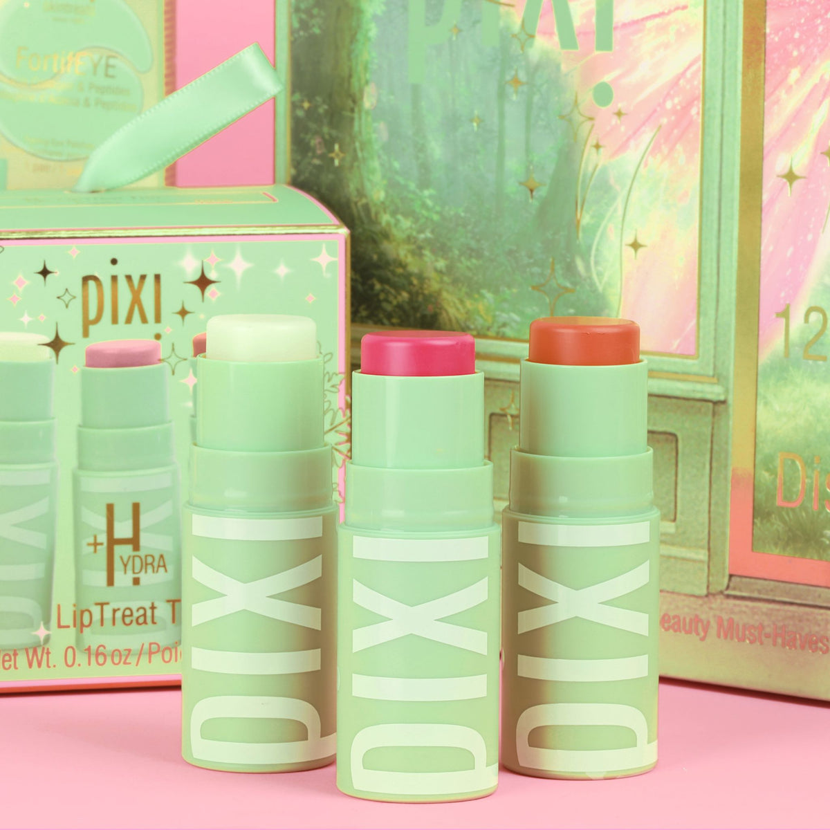 Three Pixi +Hydra liptreat lipsticks in mint green with pink, peach, and white caps, displayed in front of their colorful packaging on a pink surface.