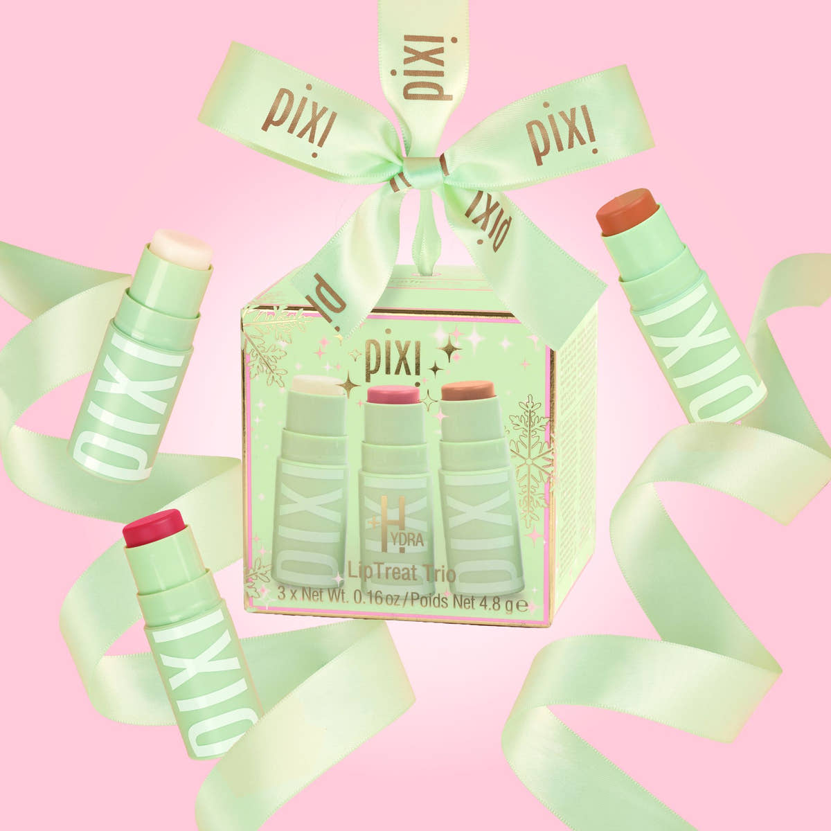 Pixi+Hydra LipTreat Trio gift set in a clear window box, wrapped in a mint green ribbon printed with the Pixi+ brand name, set against a solid pink background.